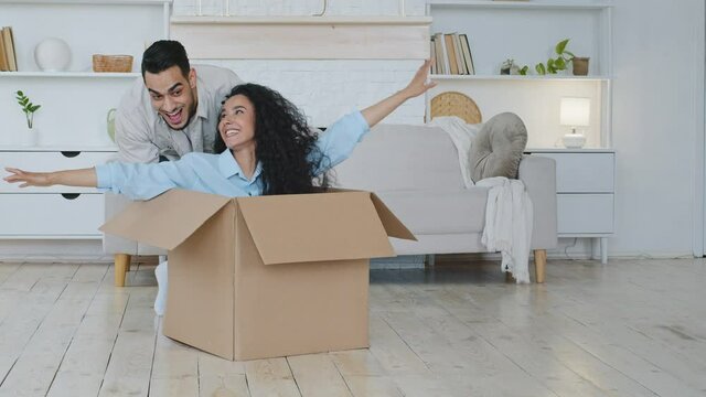 Funny curly haired lady sitting inside carton box. Hispanic man pushing parcel with woman. Happy couple renters buyers having fun packing relocate on moving day in new apartment, relocation mortgage