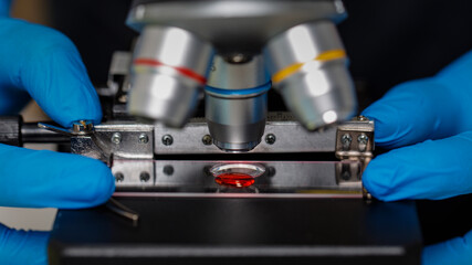 Fototapeta na wymiar Hands in blue gloves adjisting microscope with red drop of sample liquid under lens of microscope