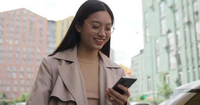 Smiling Asian Girl walking at City Street while holding Modern Smartphone. Charming Mixed-Race Young Woman in Eyeglasses enjoys Chatting using Mobile Application. Looking at Phone Screen. Social Media