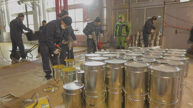 Shiny barrels with self-leveling floor workers in the background. Paint cans at a construction site. Metal drums at a construction site