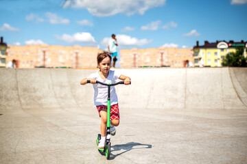 Obraz premium preschool girl riding a scooter in red shorts and a white T-shirt on the playground on an early sunny hot summer morning. active rest in the park.