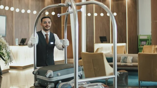 Slowmo portrait of young Middle Eastern male porter in uniform smiling to camera standing in hotel lobby holding cart with guests luggage