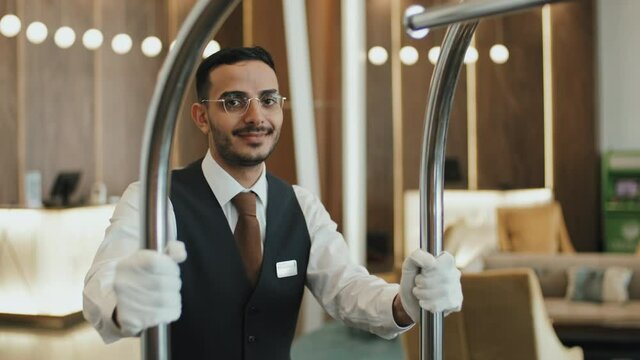 Medium slowmo portrait of smiling young male bellboy in uniform posing for camera with luggage cart standing in posh hotel lobby