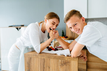 Teenage brother and her adult sister, young in 20s, arm wrestling at cozy home kitchen.