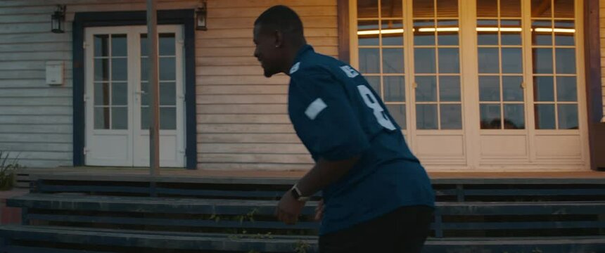 TRACKING African American father and son playing football together on a lawn in front of their house in the evening. Shot with 2x anamorphic lens