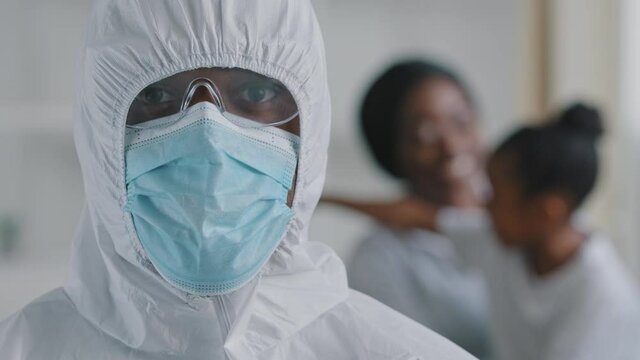 Afro american doctor scientist medical worker wears protective clothing hood transparent glasses mask looks at camera stands posing on blurred background woman with baby african mother with daughter