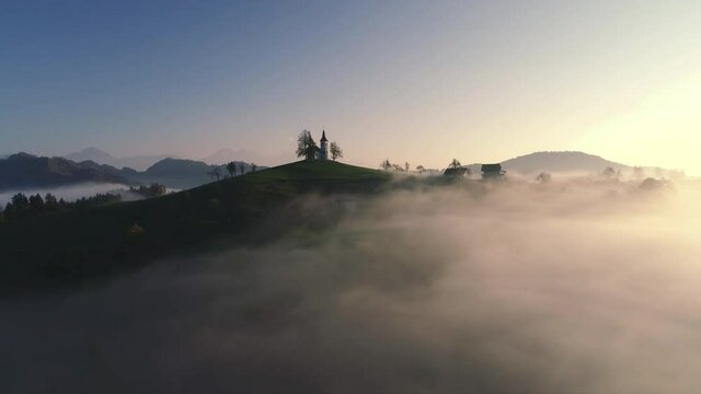 Aerial landscape of mountain hills covered in fog and Saint Thomas church, Slovenia on sunset