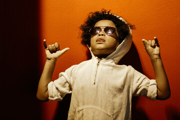 Black boy, with afro hair and sunglasses. With a cocky and funny expression like a rapper. With a red wall in the background. Children, future, education, immigration, and equality concept.