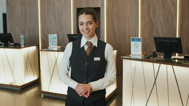 Medium slowmo portrait of young smiling female hotel manager or receptionist in uniform posing for camera standing in modern lobby with counters for check-in at background