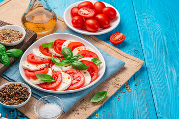 A plate of healthy classic caprese salad with tomatoes and mozzarella cheese with basil leaves on a blue background. Traditional Italian salad.