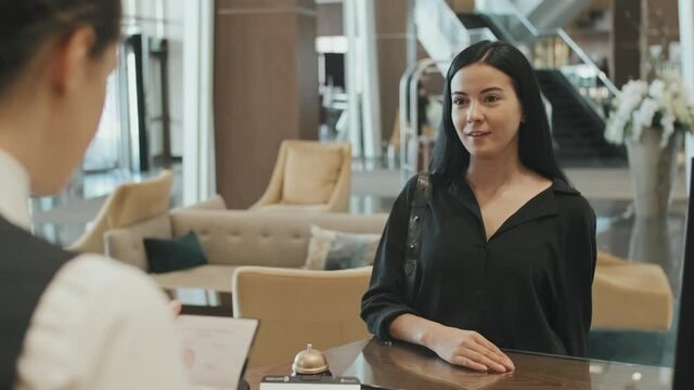 Medium shot of young attractive brunette woman approaching to reception desk at modern hotel for checking-in, giving her passport to female receptionist
