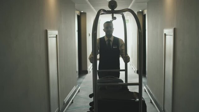 Tracking front-view shot of young male porter in uniform carrying luggage cart with guests suitcases along hotel corridor
