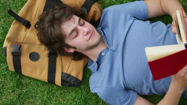 Top view of young man lying on grass reading book with head on backpack