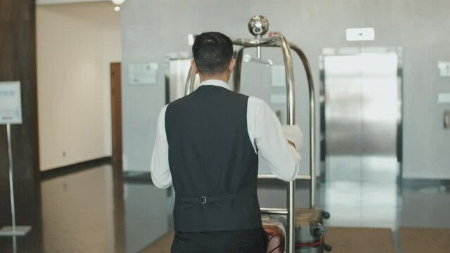 Rear-view tracking shot of male porter in uniform carrying luggage cart with suitcases along hotel lobby to elevators