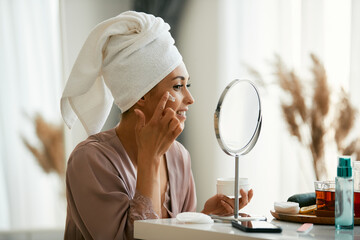 Beautiful woman applies face cream while looking in mirror at home.