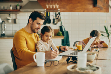 Little girl sits on father's lap while he is writing notes and working on laptop at home.