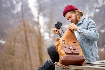 Handsome caucasian photographer male taking camera from leather handmade backpack, going to take photos. Photography, handmade accessories concept. copy space