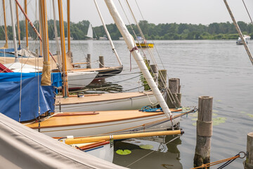 Fototapeta na wymiar Wroxham, Norfolk Broads, UK – July 2021. Sailing boats moored along the wooden staging on the side of Wroxham Broad during the annual sailing regatta open week.