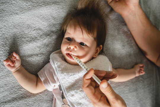 Close up on small caucasian baby four months old and hand of unknown father giving her medicine antibiotics while lying on bed at home parenthood and health issues liquid drugs oral syringe concept