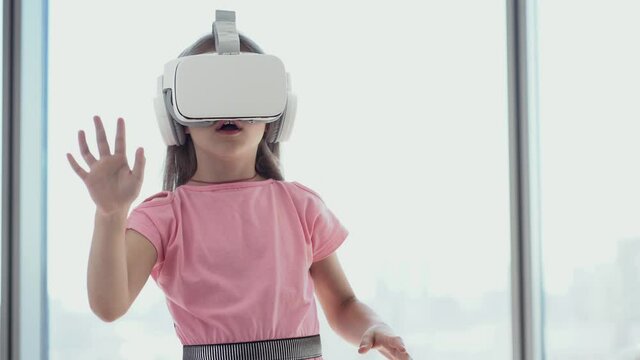 The girl is having fun in white virtual reality glasses, stands against the background of the window and touches the glasses with her hands in surprise. High quality 4k footage