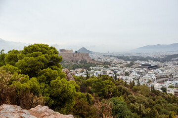 Obraz premium Acropolis (Parthenon, Temples), Mount Lycabettus in distance and white city buildings with vivid pine greenery. Athens historical landmark from Filopappou Hill on cloudy day
