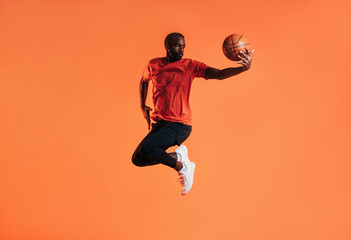 Fototapeta na wymiar Young man in sportswear practicing basketball against an orange background. African male athlete jumping in the air with basket ball in studio.