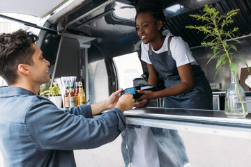 Young man with credit card paying to a saleswoman at a food truck. Female entrepreneur in apron...