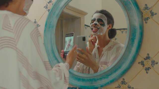 Close up of young adult African American woman wearing funny panda cosmetics facial mask recording video on mobile phone standing in bathroom. Funny 20s woman making beauty procedures at home.