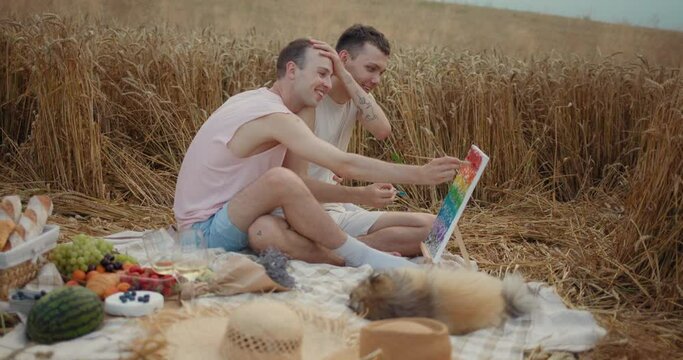 Smiling Couple of Men Gays sitting on a Picnic Blanket with Puppy and Drawing. Happy Guys Using Small Easel for Rainbow Picture while Resting at the Field. Beautiful Love Moments. Food. Wine. Males.