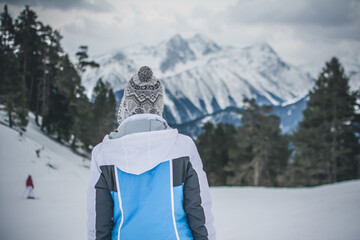 A girl stands with her back in a hat and jacket against the background of a mountain landscape