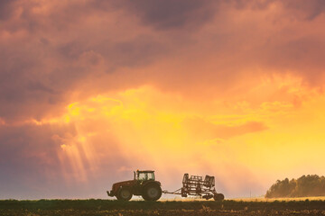 Naklejka premium Tractor On Road In Spring Season. Beginning Of Agricultural Spring Season. Cultivator Mounted On Tractor In Countryside Rural Field Landscape. Sunny Spring Sunset Sunrise Sky