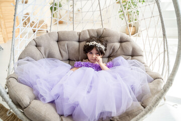 Portrait of happy brunette child girl in beautiful purple dress with pearl tiara  sitting  on swing