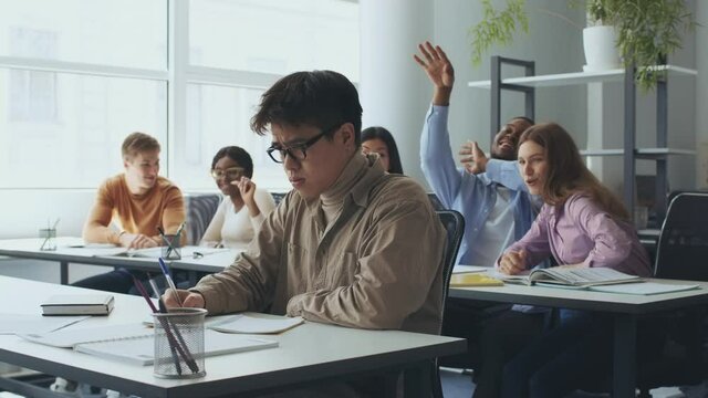Sad downtrodden asian guy nerd sitting apart from classmates, students throwing paper balls at him and laughing