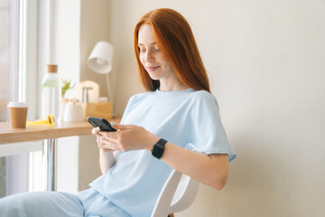Medium shot portrait of relaxed young woman in casual clothing using mobile phone sitting at desk by window in cozy light cafe. Pretty redhead Caucasian lady having leisure activity in coffee shop.