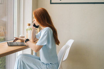 Side view of focused young woman freelancer talking on smartphone and using laptop sitting at table by window in cafe. Pretty redhead Caucasian female remote working or studying.
