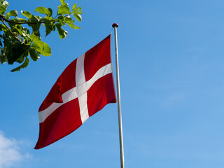 The Danish flag Dannebrog on a flagpole up against a blue sky with white clouds. Red flag with white cross. Denmark. Green leaves in forground.