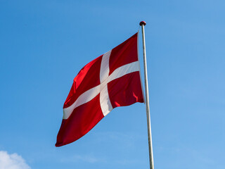 The Danish flag Dannebrog on a flagpole up against a blue sky with white clouds. Red flag with white cross. Denmark. 