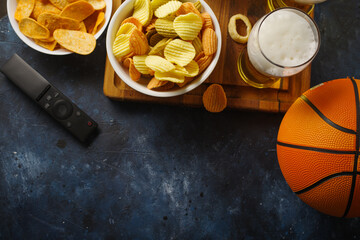 Obraz na płótnie Canvas The traditional set of a sports fan - beer, chips, snacks. There is also a remote control and a basketball ball on the table. Watching a sports match with friends, basketball.