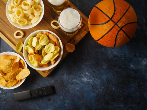 Two Glasses Of Beer, Chips, Snacks, A Remote Control And A Basketball On A Dark Blue Background. Watching A Basketball Game On TV. Rest In The Company Of Friends, Sports Fans.