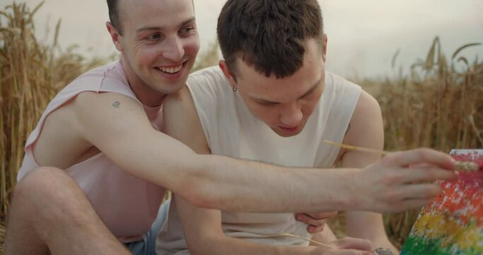 Cheerful Young Guys Lovers Drawing a Rainbow Picture while sitting in the Field. Having Good Mood and Hugging. Couple of Men Gays having a Romantic Date. Free Love. LGBT Community. Cute Males.