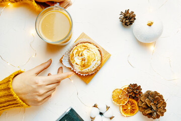 Top view of women's hands touching coffee mug and tartlet. Girl in knitted sweater is having breakfast. Sweet dessert, beverage, pine cone and decorations on white table. Still life aesthetic.