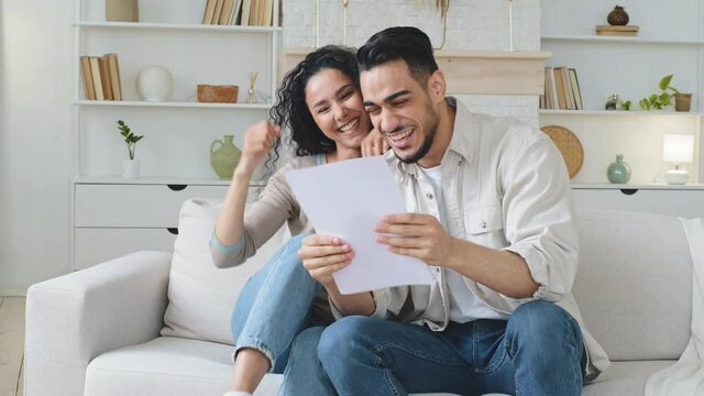Euphoric spaniard ethnic couple sit on sofa at home reading document paper letter with amazing news bank loan approval profitable job offer opportunity celebrating success hugging make victory gesture