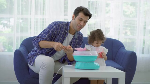 Hand father rolling cotton candy in candy floss machine and eat cotton candy together with daughter on sofa at new house