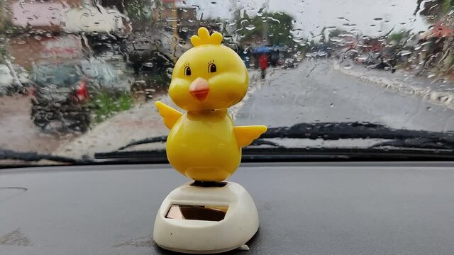 Yellow toy duck with its shaking head on top of the dashboard of a car parked on the side of the road in the rain