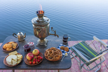 Vintage metal tea samovar with white smoke and food on the table near the calm water lake at morning