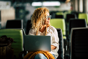 Adult blonde curly woman work at laptop computer sitting inwaiting room at the airport alone - travel and modern people in smart working remote worker activity with technology and internet - Powered by Adobe