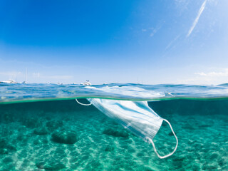 Split shot, defocused surgical face mask floating in a turquoise water with luxury yachts in the distance. Concept of sea pollution during the Covid-19 pandemic. Sardinia, Italy.