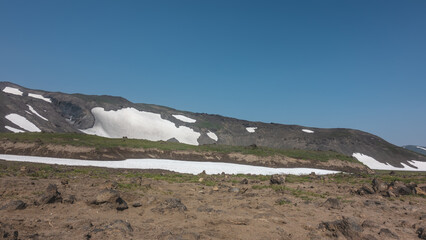 Mountaineering trail on the mountainside. There are volcanic rocks on the roadside, a little grass grows. Patches of melted snow are visible on the hill. Clear blue sky. A summer day. Kamchatka