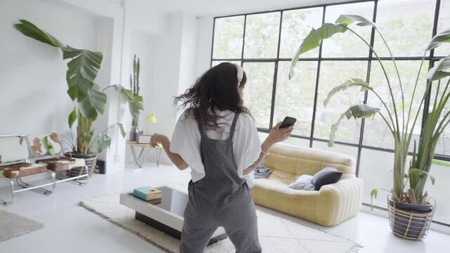 Happy carefree young girl dancing alone in a modern living room with a large window listening to music on a smartphone, funny millennial girl holding a phone enjoys success having fun at home.
