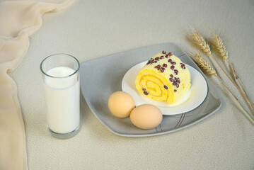 Swiss roll on a white plate, eggs, a cup of milk, a healthy breakfast on a gray background